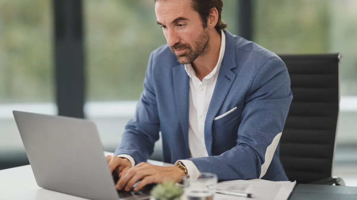 Man in blauw colbert typt op een laptop, met papieren ernaast, bij het bijhouden van overschrijvingen